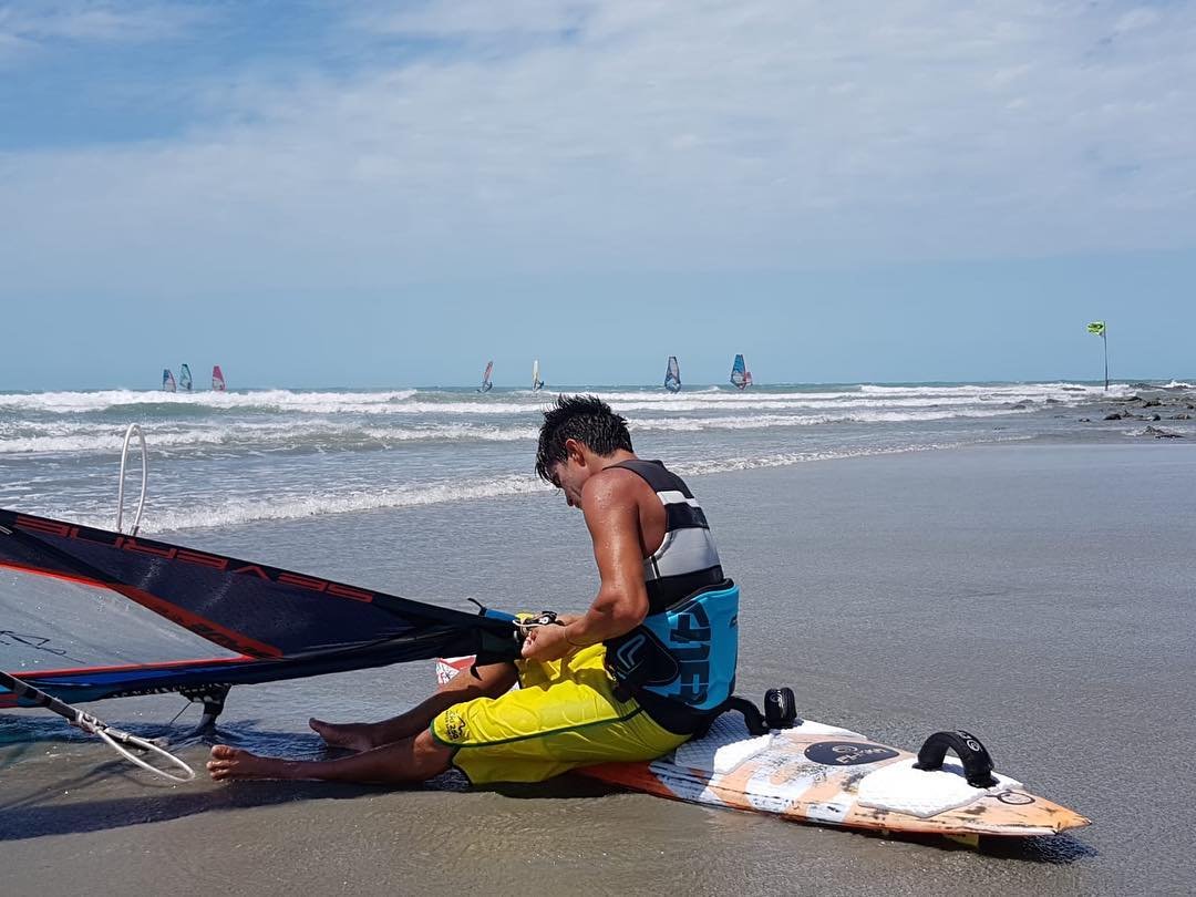 Edvan montando a vela na areia com o mar pontilhado de velas ao fundo